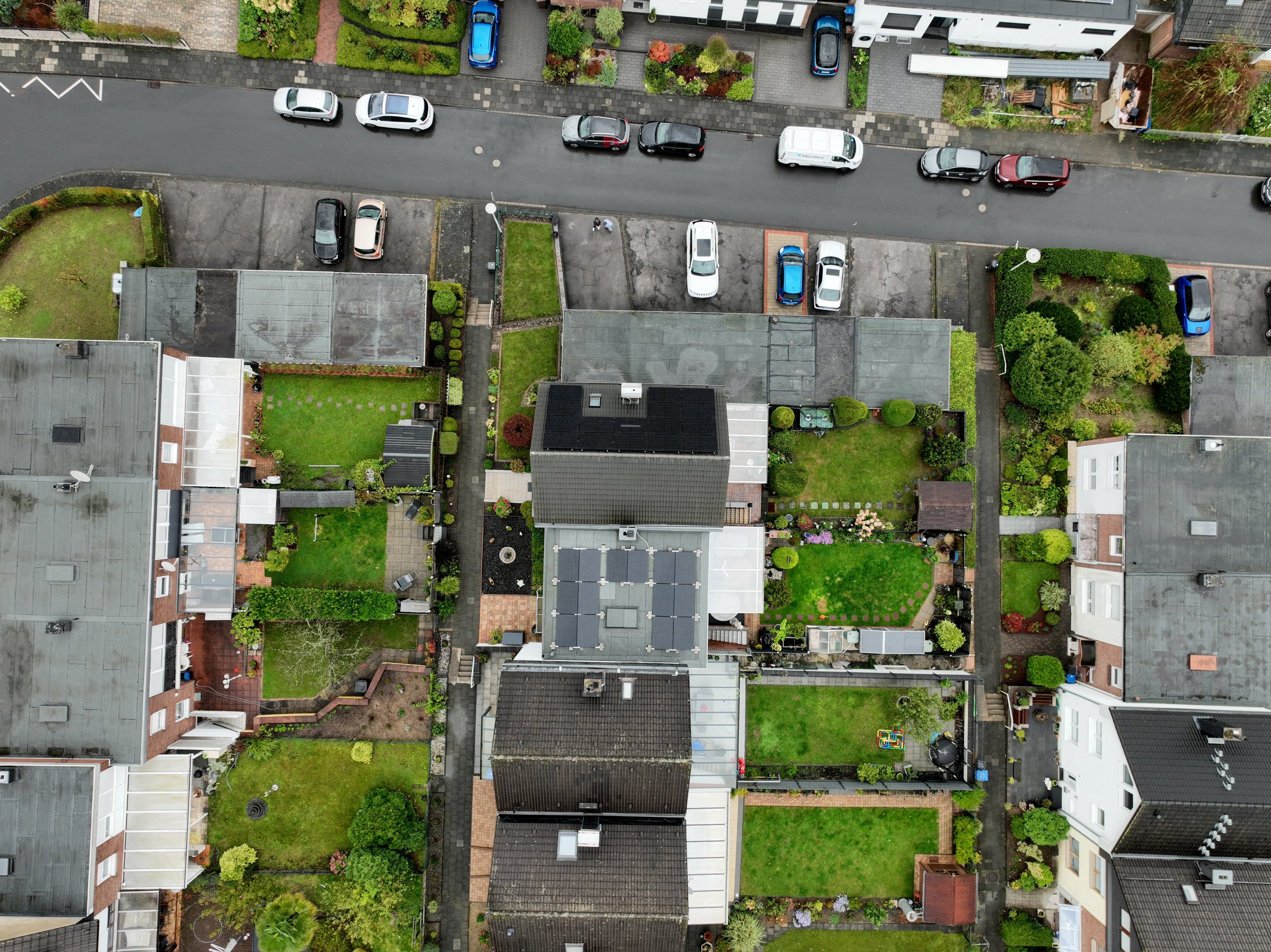 Blick von oben auf eine Wohnsiedlung mit Reihenhäusern, Gärten und geparkten Autos auf einer Straße.