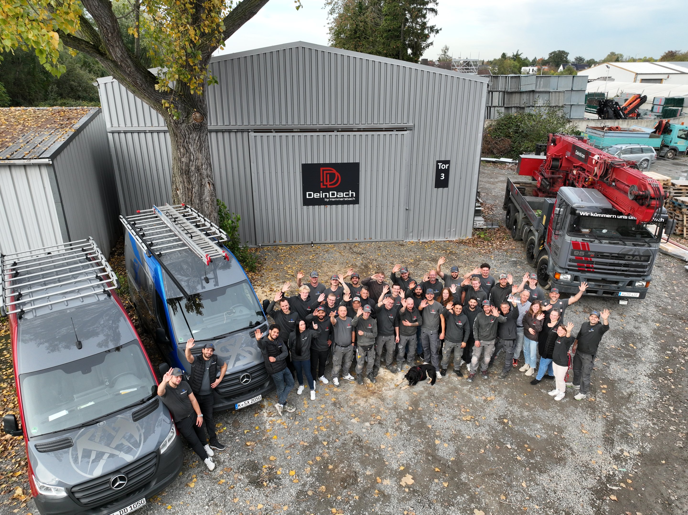Große Gruppe von Mitarbeitern der Firma „DeinDach“ in Arbeitskleidung vor einer Lagerhalle und zwei Firmenfahrzeugen.